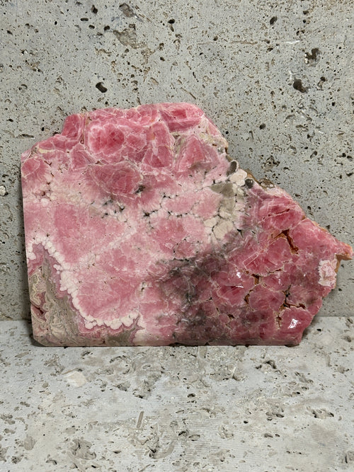 A double-polished pink Rhodochrosite mineral slab with natural growth patterns, set against a textured gray concrete background.
