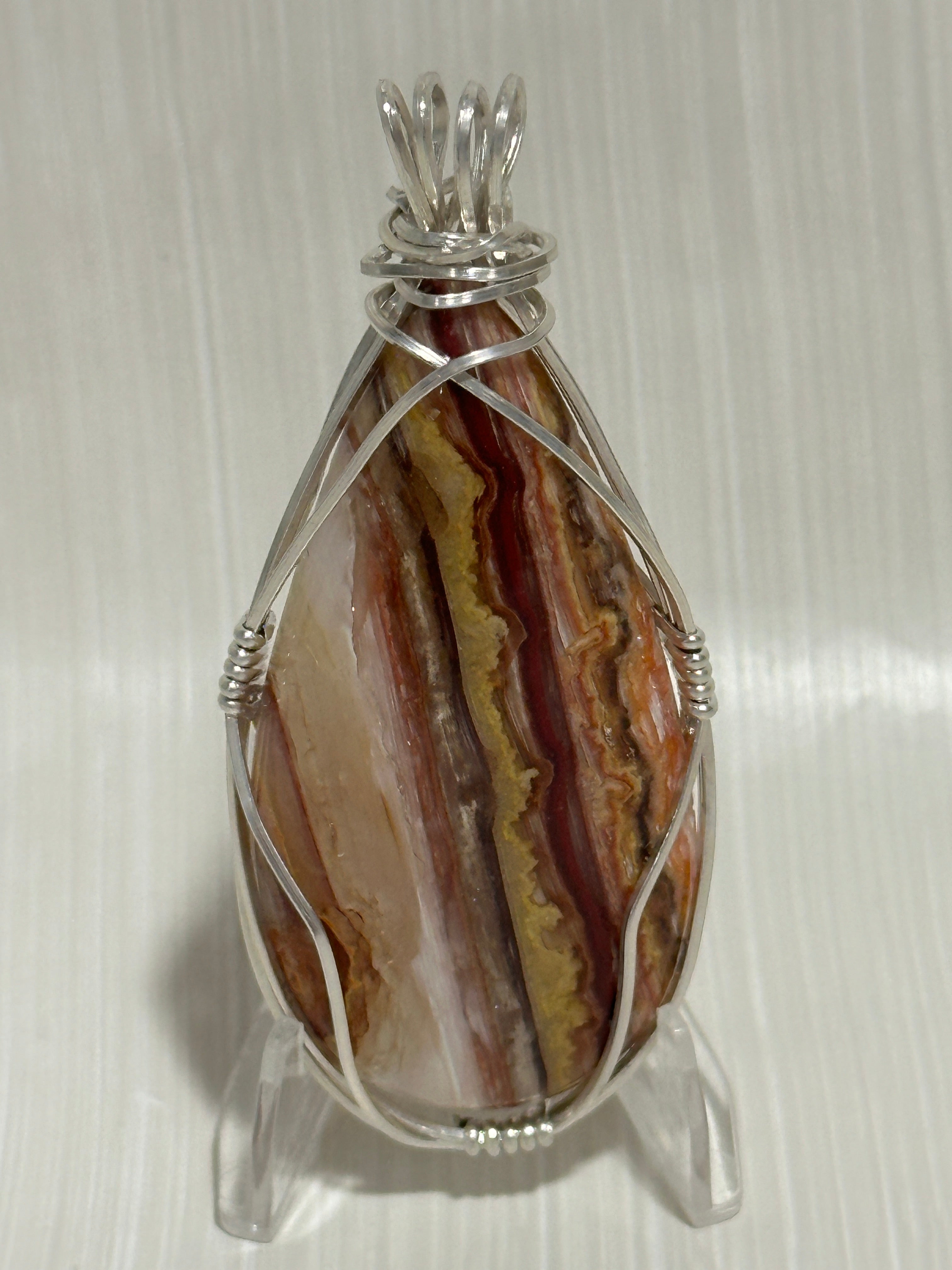 A teardrop-shaped Utah Wildfire & Candy Opal Hyalite stone pendant with banded red, brown, cream, and gold patterns, wrapped in silver wire and displayed on a clear acrylic stand.