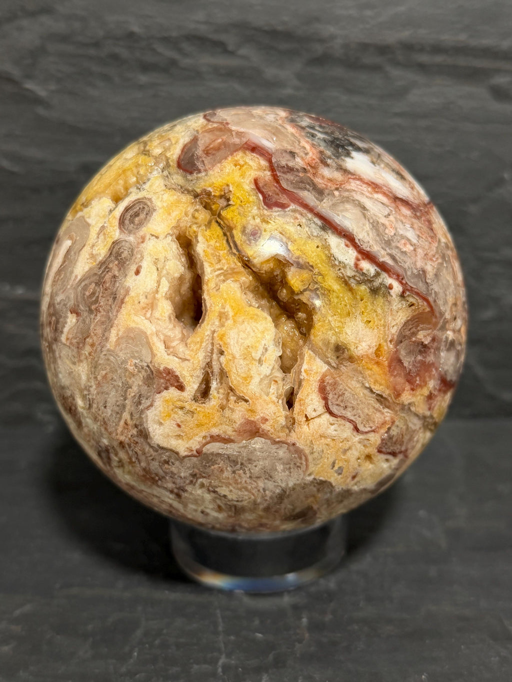 A polished decorative Utah Wildfire Bubble Hyalite stone sphere with druzy cavities and marbled patterns of beige, brown, yellow, and red, displayed on a clear acrylic stand against a dark slate background.
