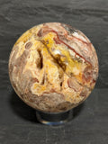 A polished decorative Utah Wildfire Bubble Hyalite stone sphere with druzy cavities and marbled patterns of beige, brown, yellow, and red, displayed on a clear acrylic stand against a dark slate background.