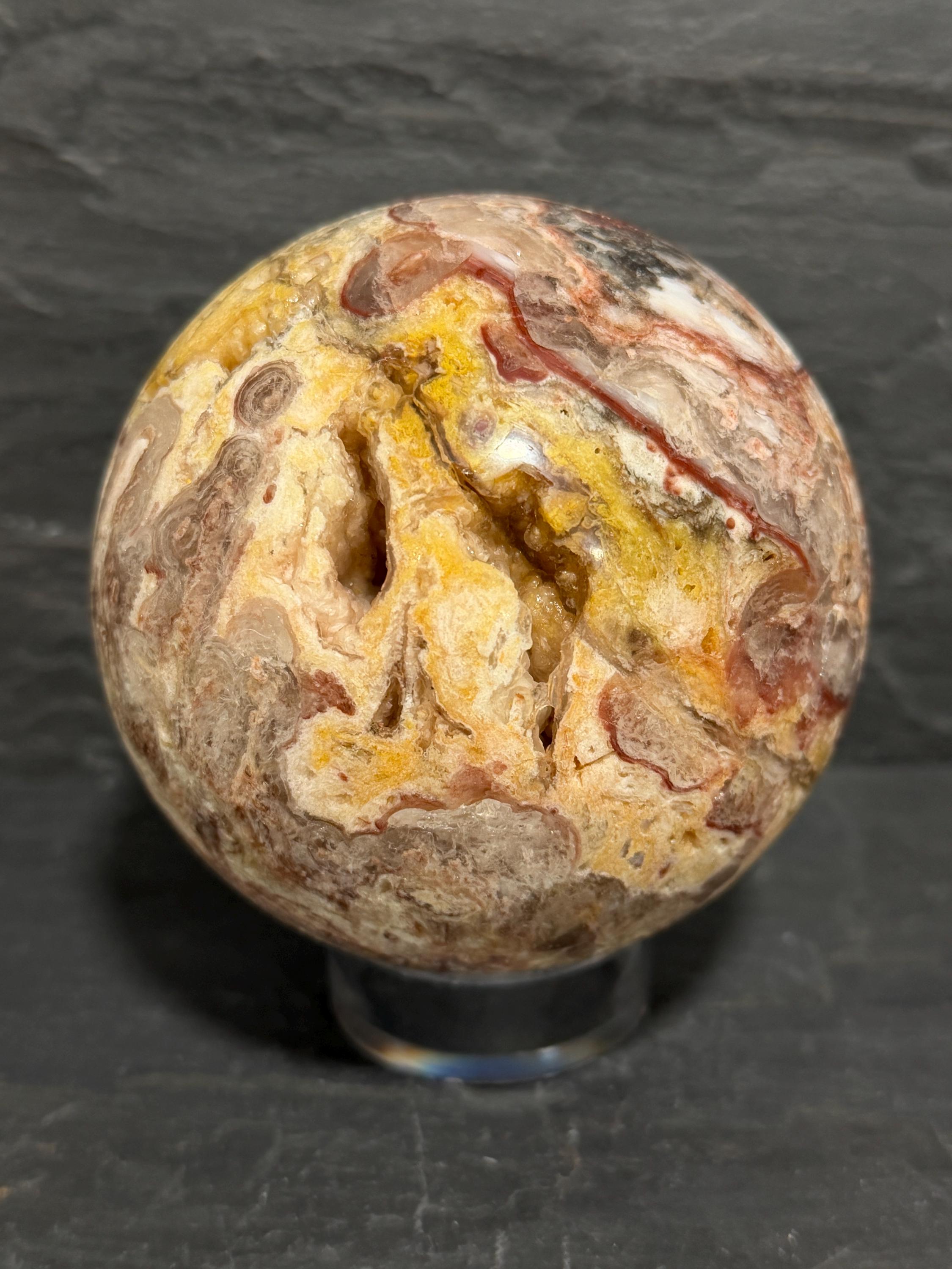 A polished decorative Utah Wildfire Bubble Hyalite stone sphere with druzy cavities and marbled patterns of beige, brown, yellow, and red, displayed on a clear acrylic stand against a dark slate background.