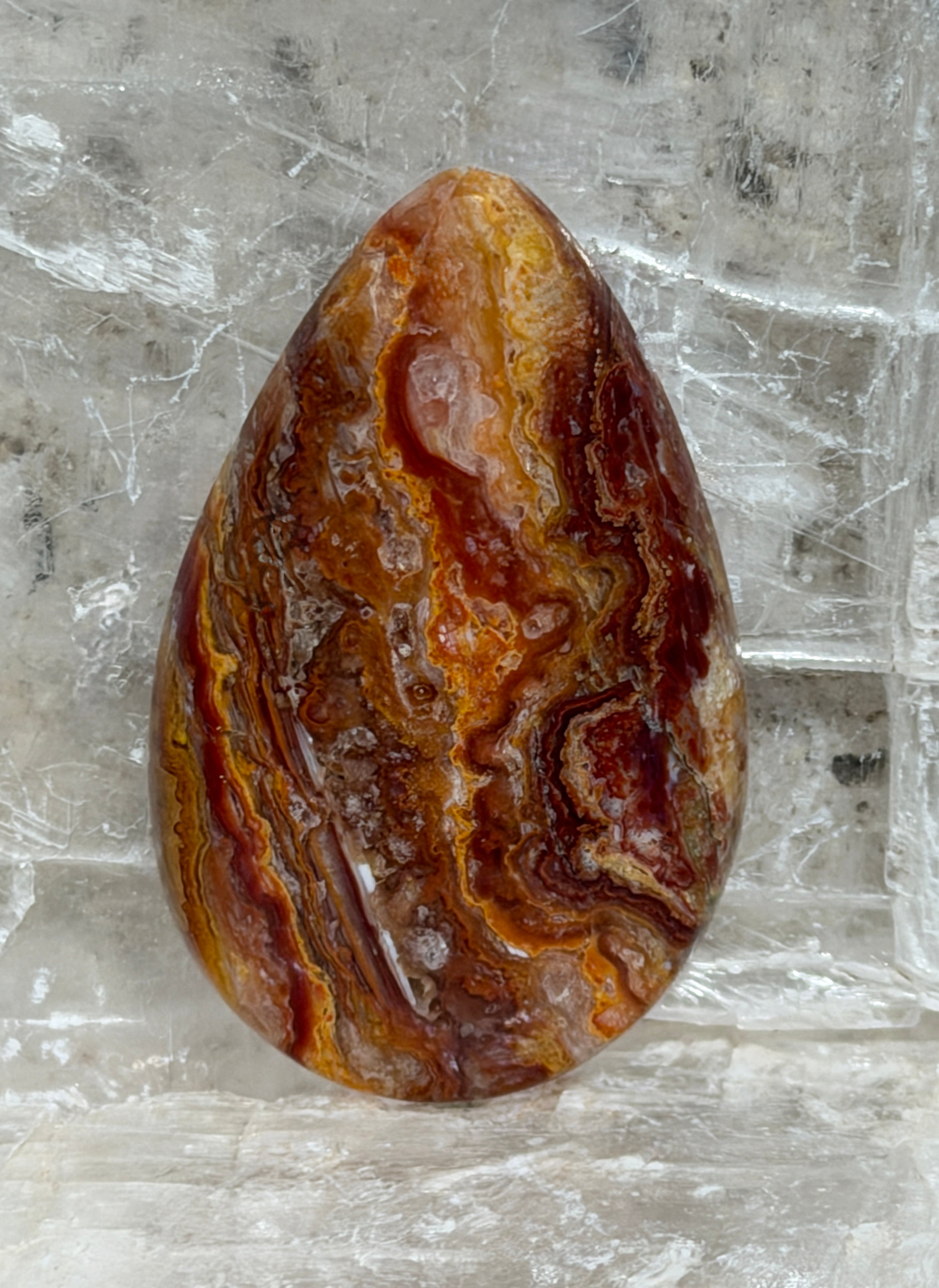 A polished Wildfire Opal Hyalite Cabochon teardrop-shaped gemstone with swirling bands of golden yellow, orange, and dark reddish-brown, resting on a clear quartz crystal cluster.